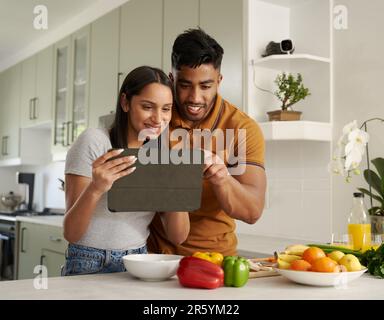 Young biracial couple in casual clothing smiling and using digital tablet by food in kitchen Stock Photo