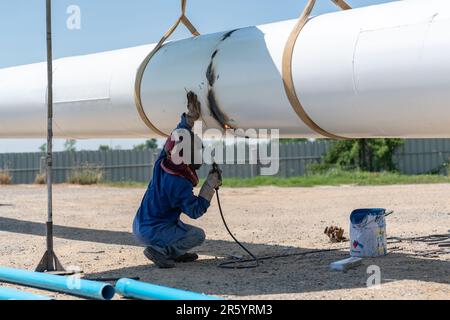 Worker in protective suit and mask crouching and welding pipe of water tower at construction site. Stock Photo