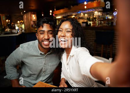 Young multiracial couple in casual clothing smiling while taking selfie with mobile phone at bar Stock Photo
