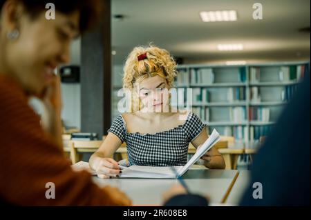 Focused woman reading book in library with crop ethnic student Stock Photo