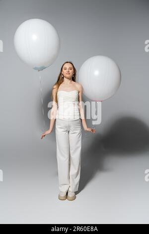 Full length of pleased and fashionable pregnant woman looking at camera while holding white balloons during gender reveal surprise party on grey backg Stock Photo