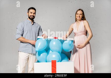 Positive pregnant woman in elegant dress opening bog gift box with blue balloons while standing near husband under confetti during baby shower on grey Stock Photo