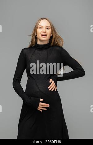 Excited hair haired and pregnant woman in stylish black dress looking at camera while standing isolated on grey, new beginnings and maternity concept, Stock Photo