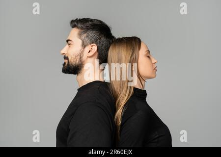 Side view of relaxed and fair haired woman standing back to back with bearded husband in black t-shirt isolated on grey, husband and wife relationship Stock Photo