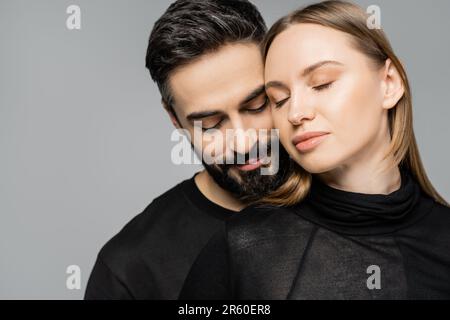 Portrait of relaxed and bearded man in black t-shirt standing next to wife with fair haired wife with natural makeup and closed eyes isolated on grey, Stock Photo
