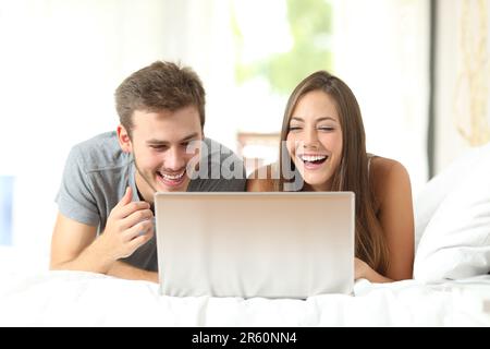 Front view portrait of a funny couple laughing watching laptop content on a bed Stock Photo