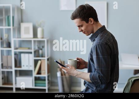 Side view portrait of smiling young man using smartphone in office at coffee break, copy space Stock Photo