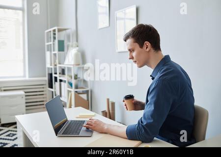 Side view portrait of young man using computer at workplace in office and drinking coffee Stock Photo
