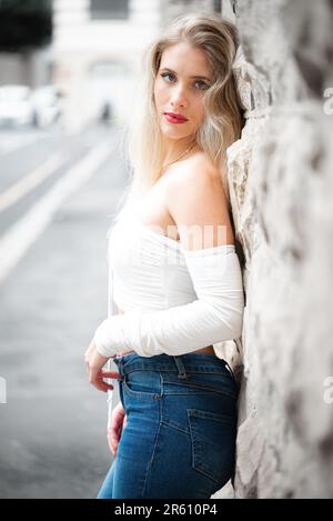 A young blonde woman in a white top and jeans posing, leaning against a wall outdoors Stock Photo