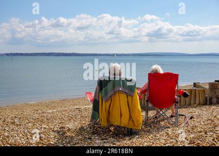 Elderly couple relaxing on stony beach. A married couple sitting on deckchairs looking out to sea on sunny day. Togetherness concept Stock Photo