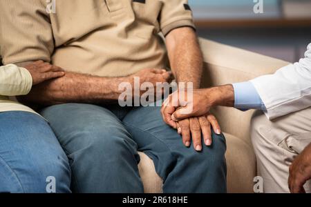 Close up shot of doctor and wife consoling senior elderly man by holding hands, while sitting on sofa at home - concept of trust, medical support and Stock Photo
