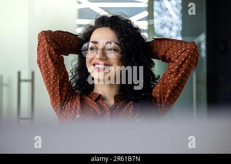 Young woman engineer developer programmer works in the office inside the IT company, satisfied with the work result, rests hispanic looks out the window, smiles hands behind her head, dreams. Stock Photo