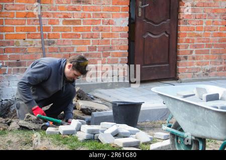 Man digging. Defocus young man laying gray concrete paving slabs in house courtyard on gravel foundation base. Master lays paving stones. Repairing Stock Photo