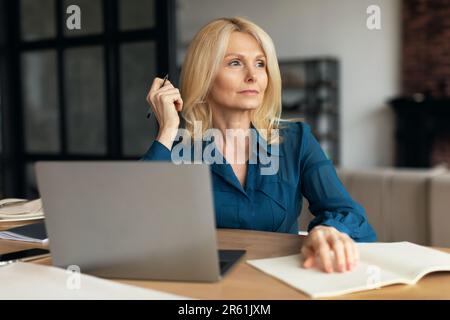 Serious pensive mature businesswoman with computer making notes, planning day, looking at free space, working at home Stock Photo