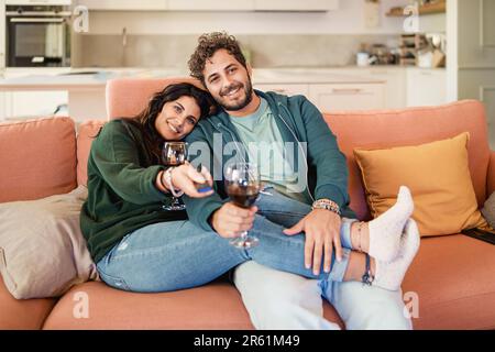 Caucasian couple, dressed casually, sitting and enjoying TV together. The woman holds the remote control while resting her feet on his lap. Relaxed an Stock Photo