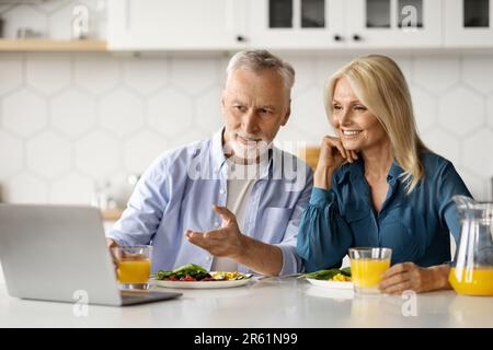 Smiling senior couple using laptop in kitchen while having breakfast together Stock Photo