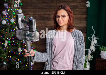 The girl is standing, smiling gently against the backdrop of the Christmas tree and holding a vacuum cleaner in her hand. Stock Photo