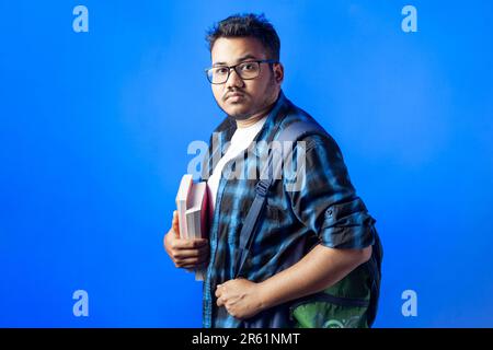 An adult college student holding books and backpack isolated in blue background. Stock Photo