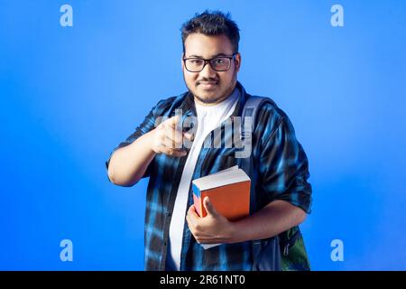 happy indian male student pointing finger towards camera in blue backdrop Stock Photo