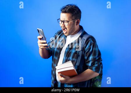 One shocked college student holding books and backpack and shouting while reading text message on his mobile phone Stock Photo