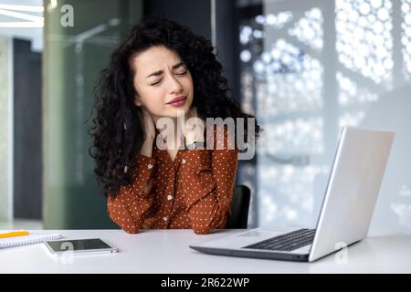 Overtired business woman working inside office with laptop, Hispanic female worker has severe neck pain, massaging muscles with hand Stock Photo