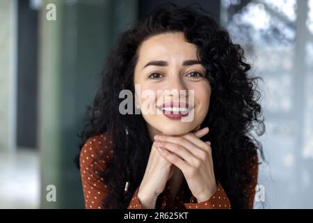 Close-up portrait of Latin American woman, businesswoman boss smiling and looking friendly at camera, female worker with curly hair successful inside office. Stock Photo