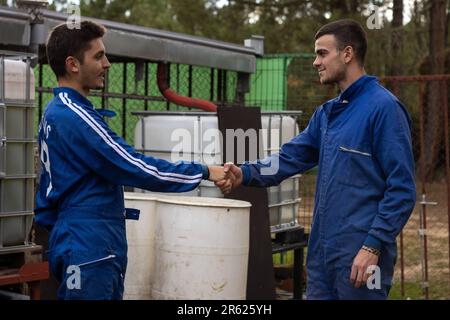 Two Caucasian men in their twenties wearing casual business attire shaking hands in a wooded area with a rustic wooden barrel and metal fence in the b Stock Photo