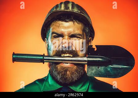 Builder in safety helmet with shovel in teeth. Male repairman, industrial worker, labourer in protective hardhat with spade. Construction worker Stock Photo