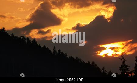 Sun setting down through clouds, above hill with coniferous trees silhouettes Stock Photo