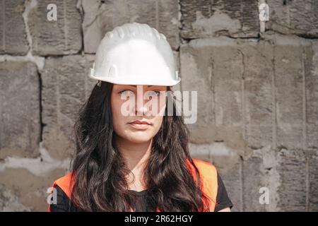 Young woman in white hard hat and orange high visibility vest, long dark hair, looking into camera. Blurred old construction site wall background Stock Photo