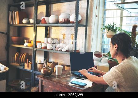 Im always an email away. an unrecognizable business owner sitting alone in her pottery studio and using her laptop. Stock Photo