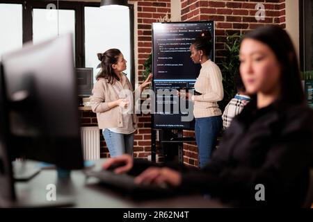 Diverse tech enginners talking about algorithms standing in front of big screen, analyzing html script language. Team of software developers discussing database on terminal window in it agency Stock Photo