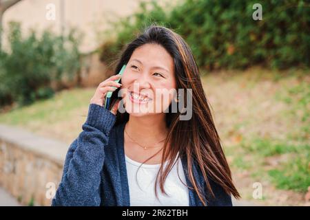 Joyful teenage female student smiling and having a cellphone call conversation. Happy carefree young chinese woman talking by smartphone outside. Asian gossip lady using a mobile phone to communicate. High quality photo Stock Photo