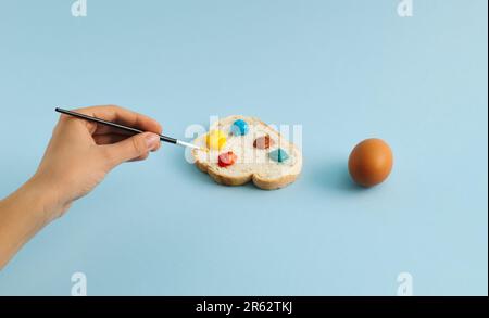 An boiled egg in hand and slice of bread with colorful palette on pastel blue background. Woman is coloring eggs for Easter holiday. Creative Easter c Stock Photo