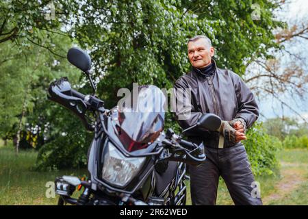 Portrait of senior biker taking off gloves by motobike outdoors. Man wearing leather jacket and trousers. Traveling alone. Summer trip Stock Photo