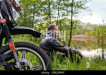 Portrait of senior biker relaxing by motobike outdoors. Man wearing leather jacket enjoying river landscape. Elderly motocyclist traveling suring summ Stock Photo
