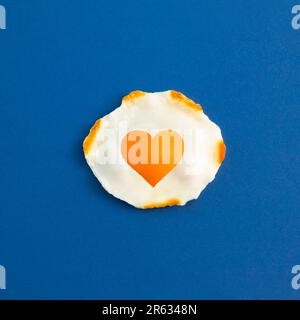 Creative idea with fried egg in heart shape on a dark blue background. Minimal food and love concept. Breakfast idea. Flat lay, top view. Stock Photo
