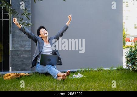 A woman sits on the grass with a laptop and rejoices raising her hands up. Stock Photo