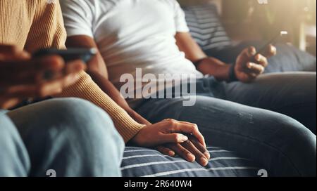 Striking connections in their own way. Closeup shot of an unrecognisable couple holding hands while using their cellphones at home. Stock Photo