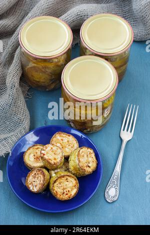Fried zucchini slices pickled in olive oil with herbs and filled in a canning jar. Vertical. Blue plate. Stock Photo