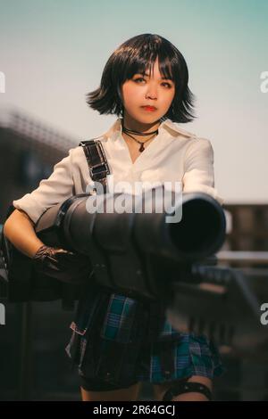 A young Asian woman stands outdoors in a white shirt and patterned dress, holding a large rifle Stock Photo