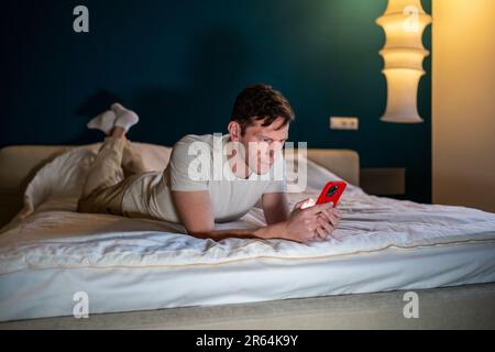 Man reading information in smartphone lying in bed at home in modern bedroom in evening. Stock Photo