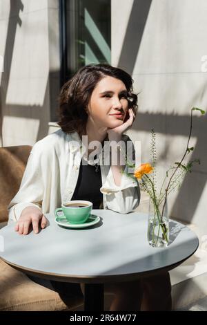 smiling woman sitting at white round table near cup of black aromatic coffee, saucer, glass vase with yellow rose and green plants on summer terrace o Stock Photo