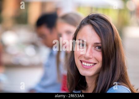 Positive young female in casual clothes and with long hair smiling and looking at camera while standing in daylight on city street against blurred bac Stock Photo