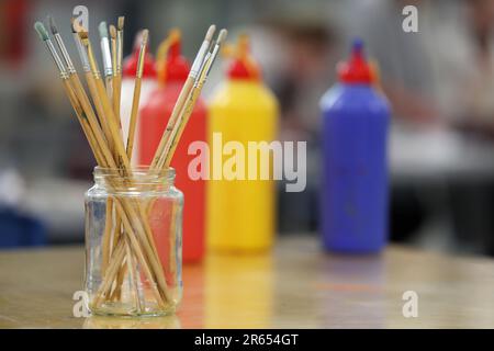 A close up of a visual art classroom with a set of paint brushes standing up in a glass jar. Primary colors of red, yellow and blue behind. Stock Photo