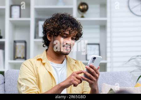 A young Hispanic man is sitting at home on the sofa in the living room, the man is holding a phone in his hands, using an online application on a smartphone, browsing the Internet and typing messages. Stock Photo
