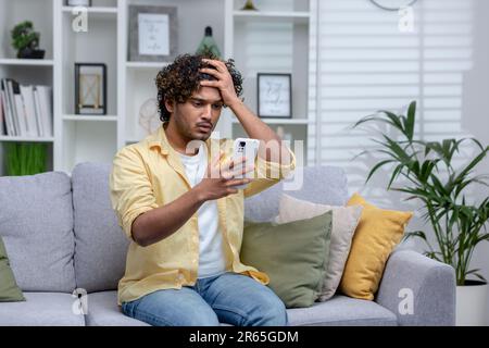 Latin american man sitting on sofa at home in living room, man upset and depressed reading bad news online using app on phone, holding smartphone. Stock Photo