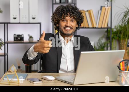 Like. Happy young indian businessman guy working on laptop looking approvingly at camera showing thumbs up, like sign positive something good, positive feedback at office workplace. Freelancer man Stock Photo