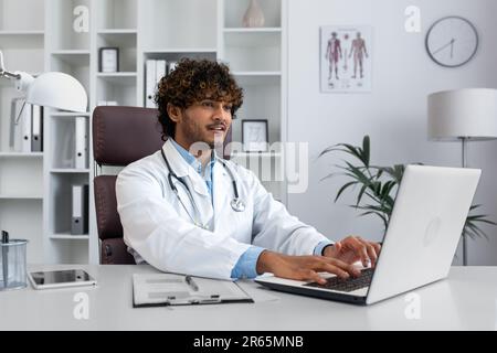 Young successful doctor working inside hospital office, man smiling and looking at laptop screen, hispanic man in medical gown is satisfied with patient's treatment achievement results. Stock Photo