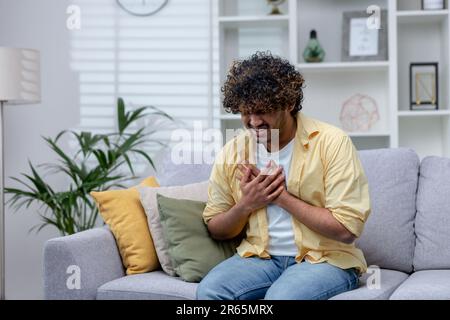Sick man having severe chest pain and heart attack, hispanic young man sitting alone at home in living room, holding hands to chest. Stock Photo